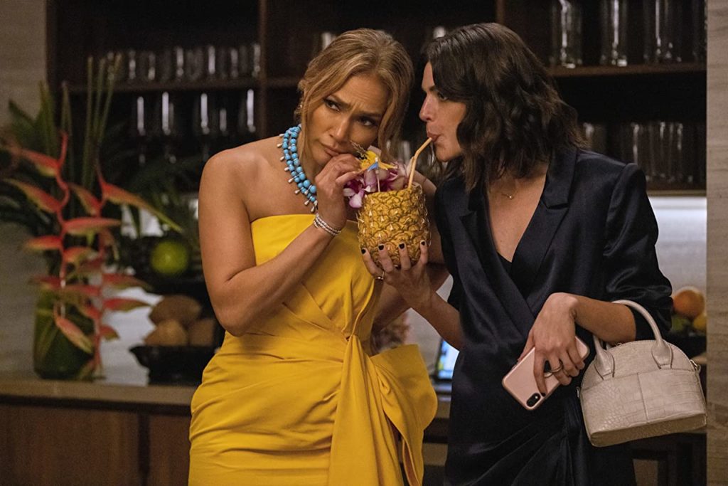 Jennifer Lopez and Callie Hernandez in Shotgun Wedding. They're both drinking a cocktail served in a pineapple out of straws and looking quizzically at something off-screen.