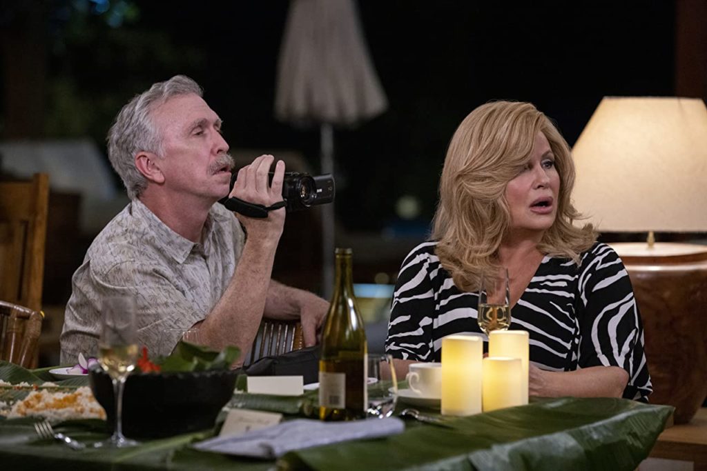 Steve Coulter and Jennifer Coolidge in Shotgun Wedding. They're sitting together at a table and look surprised at something happening off-screen. He's filming whatever's happening with an old hand-held camcorder.