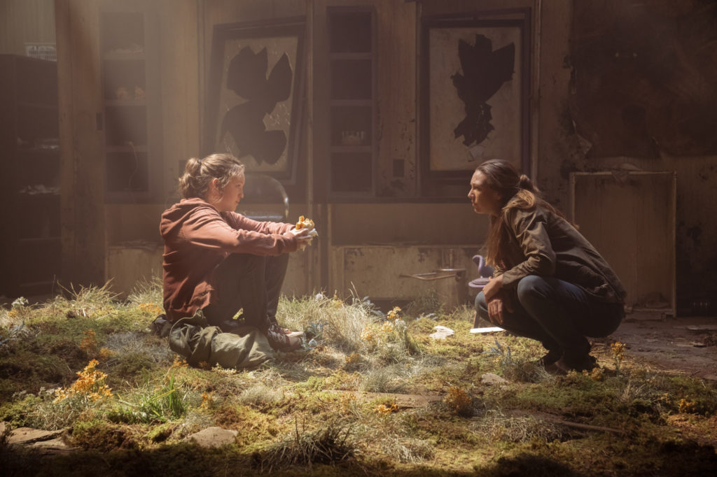 Bella Ramsey as Ellie and Anna Torv as Tess in The Last of Us. Ellie's sitting on the ground of an abandoned building while Tess kneels in front of her to speak.