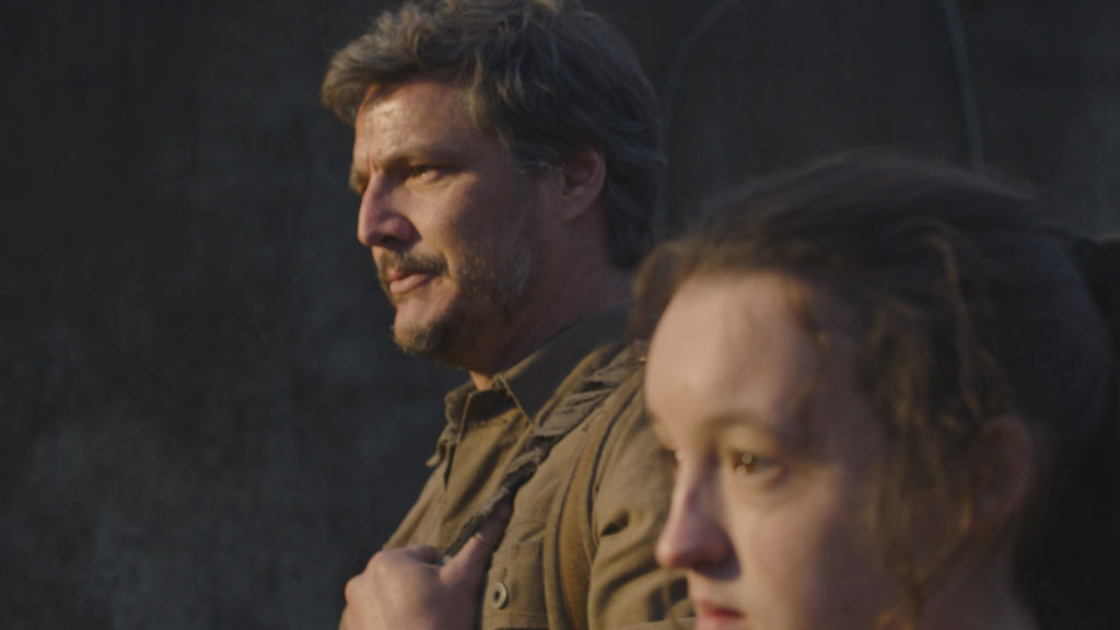 Pedro Pascal as Joel and Bella Ramsey as Ellie in The Last of Us. They're standing next to each other looking at something off camera.